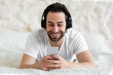 Handsome guy using smartphone and headset, bedroom interior