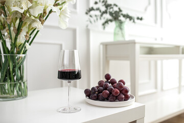 Glass of red wine with grapes and vase on white table