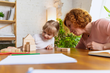 Smiling woman and cute little girl, mother and daughter playing with wooden toy cubes at home. Concept of family, support, studying, childhood, emotions