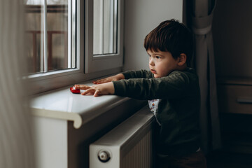 Little, pensive, two years old boy sitting by the window and playing with toy children's car
