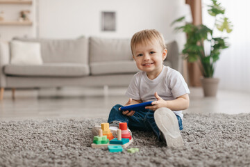 Portrait of adorable toddler boy holding smartphone, sitting on floor carpet, looking and smiling at camera, copy space