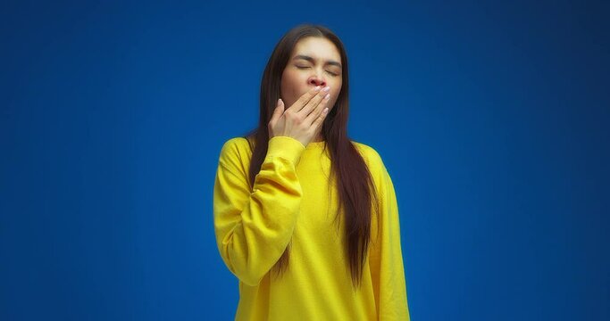 I want to sleep. Tired young woman dressed in a yellow jumper stands on a blue background in the studio, yawns, closes her eyes and makes a gesture of sleep.