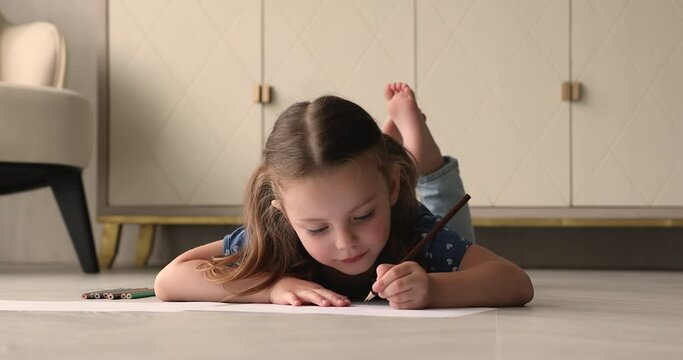 Cute small baby girl lying on warm heated floor, drawing in paper album with colored pencils. Funny little child involved in painting, spending leisure hobby pastime alone in modern living room.