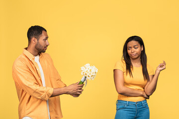 Angry black girlfriend rejecting flowers and apologies of her sad boyfriend after fight on yellow studio background