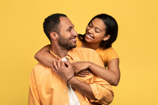 Affectionate Young Black Woman Hugging Her Boyfriend, Looking In His Eyes On Yellow Studio Background