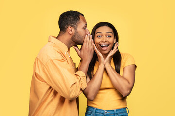 Young black guy sharing secret or whispering gossip into his girlfriend's ear on yellow studio...
