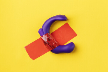 Silicone vibrator purple on yellow background. Sex toy is glued with red tape to yellow wall.