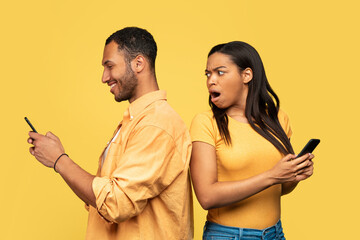 Shocked black woman spying on her boyfriend who is using smartphone, chatting with lover, cheating on yellow background