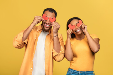 Blinded by love concept. Affectionate African American couple covering eyes with red paper hearts on yellow background