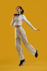 Fototapeta na wymiar Inspired graceful lady dancing, with headphones around neck in studio. Front view of happy young girl in casual clothes, jumping with earphones, isolated on orange background. Concept of inspiration.