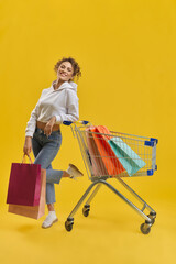 Pretty girl with bags, standing, shyly bending one leg on knee in studio. Front view of female...