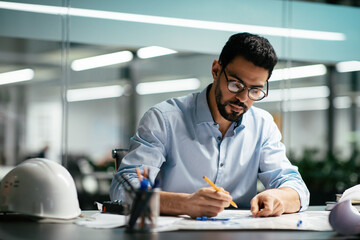 Serious attractive millennial arabic man engineer in glasses with beard works with drawings at...