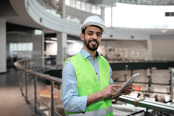 Happy millennial muslim male engineer in protective uniform and hard hat with beard and tablet in...