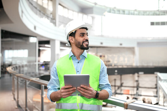Cheerful millennial muslim engineer guy in protective uniform, hard hat with beard hold tablet, look at empty space