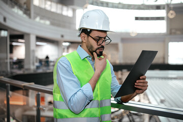 Pensive millennial muslim guy engineer in protective uniform and hard hat in glasses with beard looks at tablet