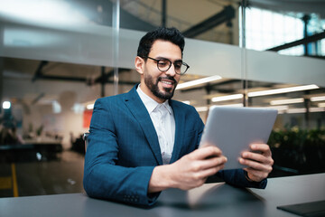 Fototapeta na wymiar Smiling busy handsome millennial middle eastern businessman with beard in glasses, suit works with tablet