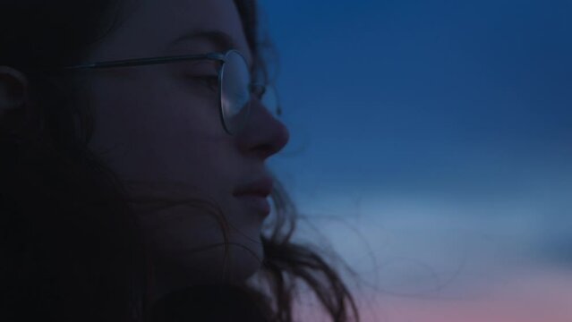 Closeup cinematic soft side view portrait of pretty young woman or teen with dark curly hair wearing casual eyeglasses, dreaming, thinking, relaxing outdoors on sunset weekend, trip, travel to Porto