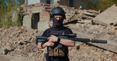 A soldier with a machine gun stands against the background of a destroyed house, looks around and raises his weapon.