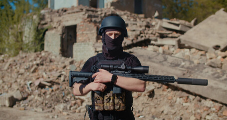 Fototapeta premium A soldier with a machine gun stands against the background of a destroyed house, looks around and raises his weapon.