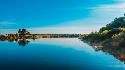 Fototapeta premium Aerial view of a beautiful summer landscape over river while dawn. Top view over river with a smooth water surface reflecting blue sky.