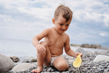 Cute Caucasian boy playing on a pebble beach with a plastic toy. Active holidays on the coast