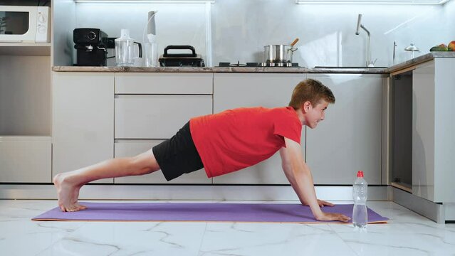 Skinny teen boy goes in for sports in quarantine in kitchen on yoga mat. Guy does push-up exercise through strength and lies on floor from exhaustion but is satisfied. It is difficult to train