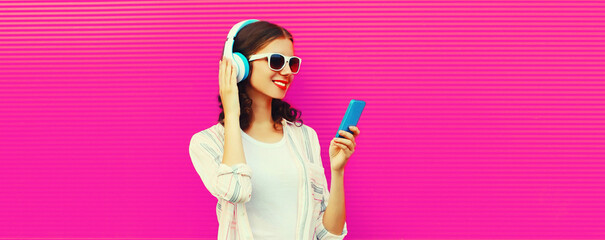 Portrait of happy smiling young woman in headphones listening to music with smartphone on pink background, blank copy space for advertising text