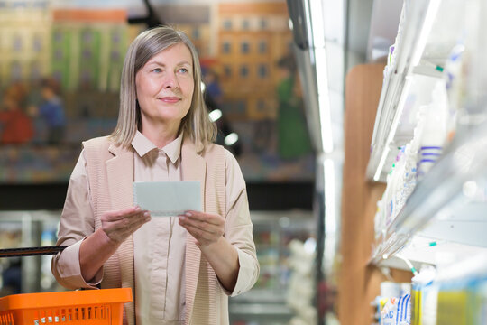 Senior woman in the store chooses goods in the dairy department near the refrigerators