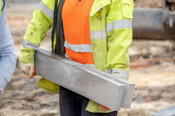 Close up of groundworker in orange and yellow hi-viz  carrying heavy concrete kerbs on construction...