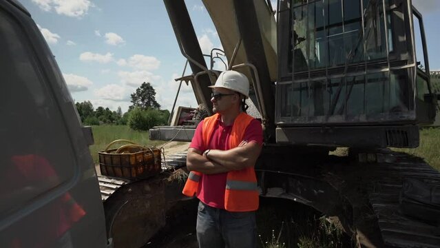 Chief builder in sunglasses retroreflective vest and construction helmet standing near excavator and controls working process