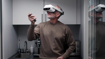 Mature man using augmented reality headset and joystick standing in small kitchen