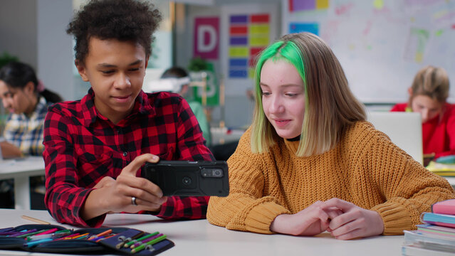 Diverse teenage classmates watch video on smartphone sitting at desk in classroom