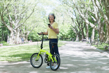 Obraz premium Senior beautiful woman walks in the park, rides a bike, listens to music on headphones, holds a phone. He stands with a bicycle in his hands, looks at the camera, smiles.