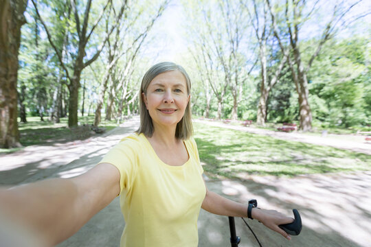 Senior gray-haired woman, active sportswoman with yellow bike in the park in the summer looks at the phone camera and smiles waving her hand at a video call near office center