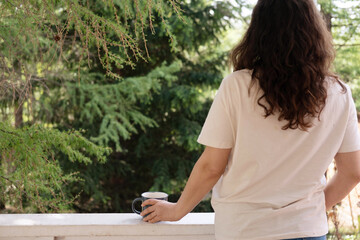 A romantic portrait of a beautiful young woman in a T-shirt and jeans, resting on a balcony overlooking the forest, with a cup of tea or coffee in her hands.