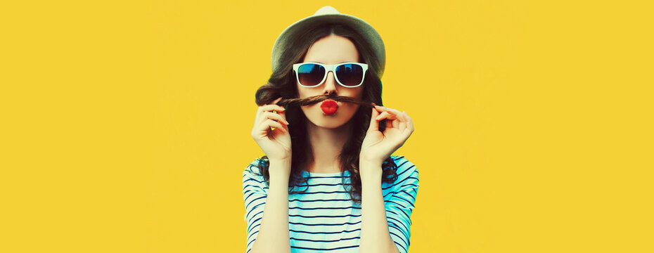 Portrait of funny young woman showing mustache her hair blowing lips with red lipstick sending air kiss on colorful yellow background, blank copy space for advertising text