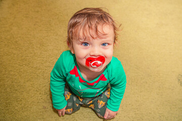 cheerful smiling red-haired baby boy sits on the floor with a pacifier in his mouth