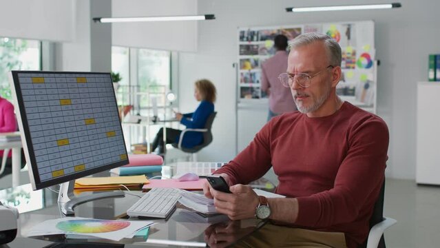 Mature businessman sitting at desk and chatting in smartphone in creative office