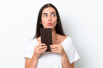 Young caucasian woman isolated on white background taking a chocolate tablet and having doubts