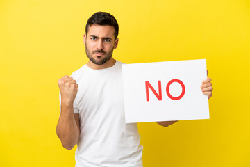 Young handsome caucasian man isolated on yellow background holding a placard with text NO and angry