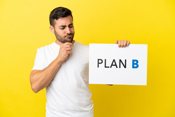 Young handsome caucasian man isolated on yellow background holding a placard with the message PLAN B and thinking