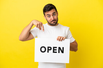 Young handsome caucasian man isolated on yellow background holding a placard with text OPEN and  pointing it