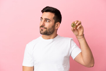 Young man holding a Bitcoin isolated on pink background looking to the side