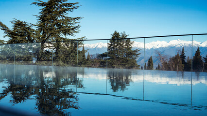 Outdoor swimming pool at the resort. Mountains landscape panorama. Snow on the tops of the mountain range. Reflection of trees and mountain peaks on the surface of blue water. Sunny day in travel