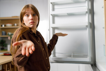 Surprised woman shrugs shoulders near empty refrigerator