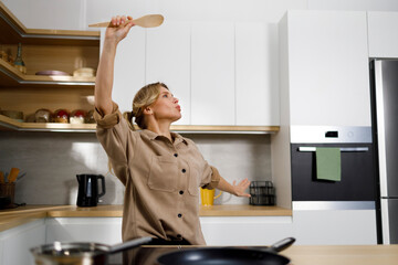 Playful woman dancing with wooden spatula while cooking in home kitchen