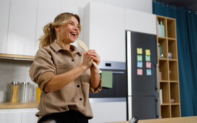 Pretty woman with joyful mood sings in the kitchen while cooking using a wooden spatula as a microphone