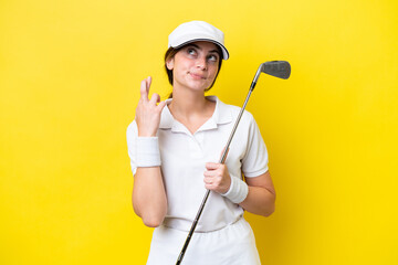 young caucasian woman playing golf isolated on yellow background with fingers crossing and wishing...