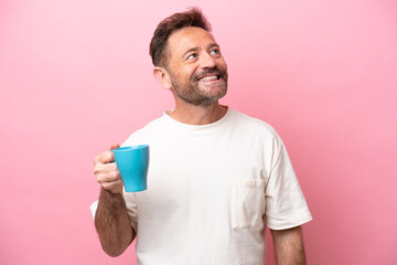 Middle age caucasian man holding cup of coffee isolated on pink background looking up while smiling