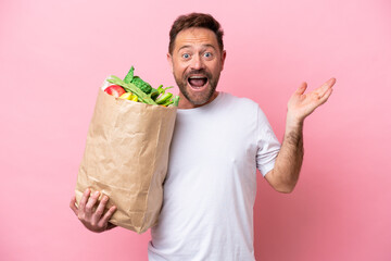 Middle age man holding a grocery shopping bag isolated on pink background with shocked facial...